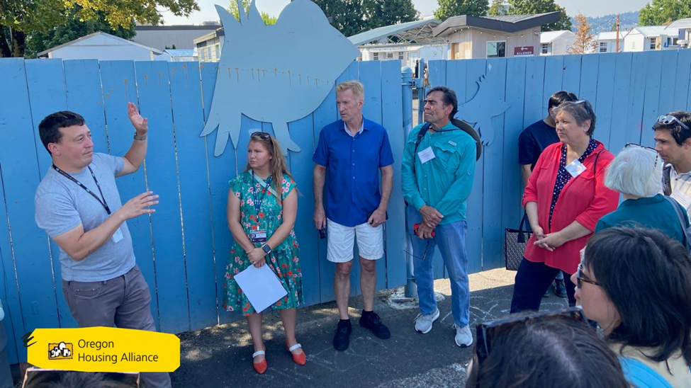 group of people listen to a man speak with a blue fence and some small structures peaking over the top