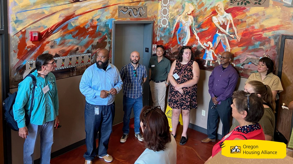 group of people listening to a man speak with a backdrop of a colorful abstract mural